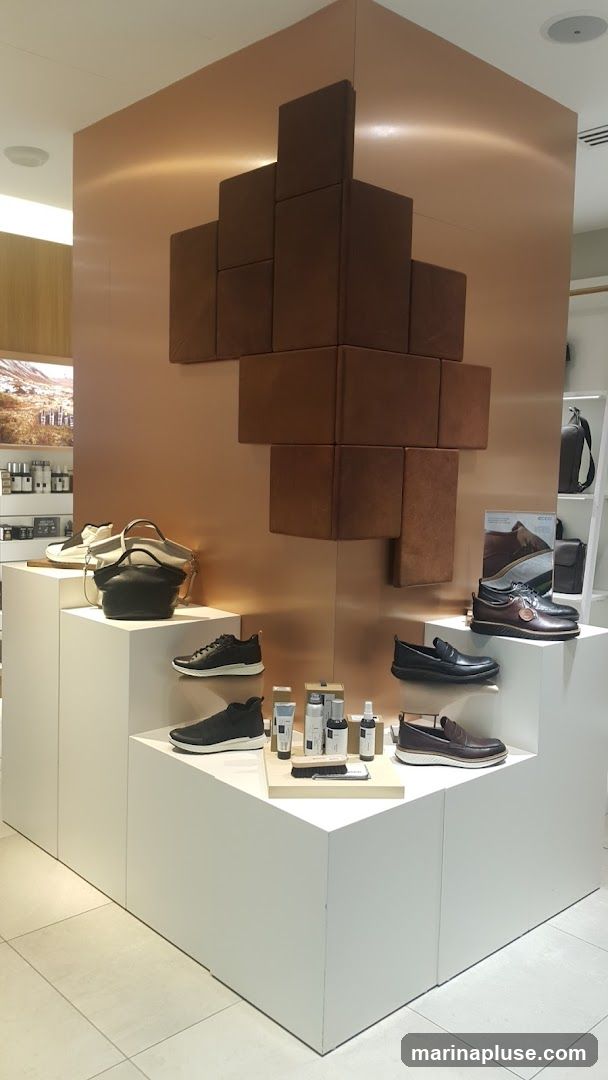 A retail display featuring various shoes and handbags on white tiered platforms. A copper-colored wall with a geometric brown panel design serves as the backdrop. Several bottles of shoe care products are also displayed.