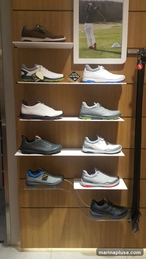 A display of various golf shoes on white shelves against a wood-paneled wall. A framed picture of a golfer is on the upper right. Several shoes have GORE-TEX tags.