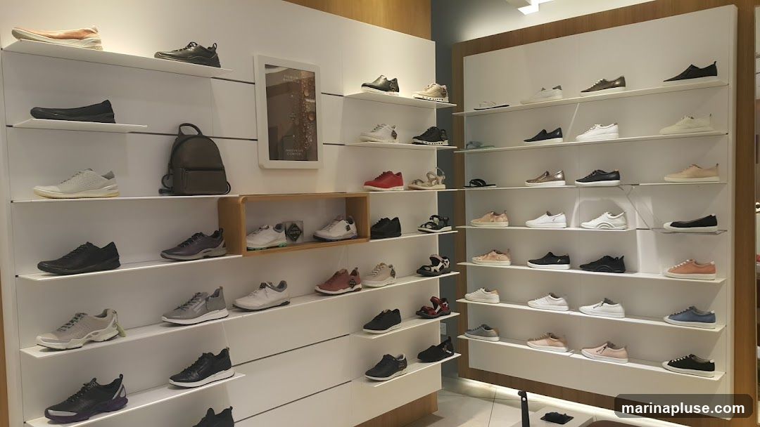 A well-lit shoe store interior with multiple shelves displaying a variety of footwear for men, women, and children. The shelves are organized by shoe type and color, with sneakers, loafers, sandals, and dress shoes visible. A dark gray backpack is also displayed on one of the shelves. A framed advertisement or informational poster is mounted on the wall.