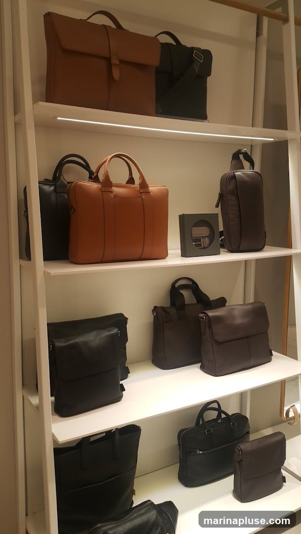 A retail display featuring various leather bags on white shelves. The bags include briefcases, messenger bags, and crossbody bags in brown and black.