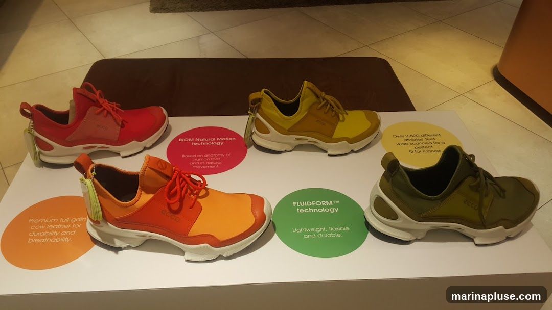 A display of four Ecco brand shoes in different colors: red, yellow, orange, and olive green. Each shoe is placed on a white surface with informational circles. One circle describes 'BIOM Natural Motion technology' and states it's 'Based on anatomy of human foot and its natural movement.' Another circle highlights 'FLUIDFORM™ technology' and describes it as 'Lightweight, flexible and durable.' A third circle mentions 'Premium full-grain cow leather for durability and breathability.' A fourth circle states 'Over 2,500 different athletes feet were scanned for a perfect fit for runners.'