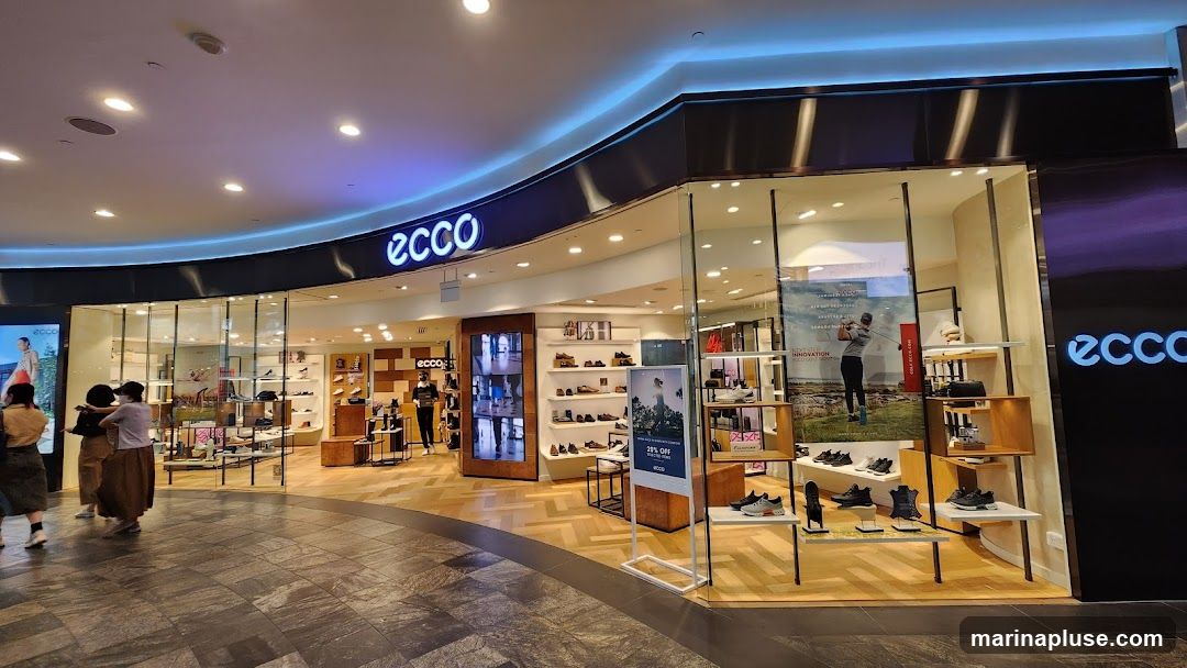 The exterior of an Ecco shoe store, featuring a prominent 'ecco' logo above the entrance. The store has large glass windows displaying various shoes on shelves. Inside, customers can be seen browsing the merchandise. The flooring is a patterned wood, and the ceiling has modern lighting.