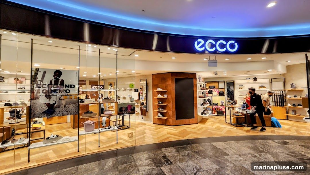 The exterior of an Ecco shoe store with a large glass window displaying shoes and accessories. The Ecco logo is prominently displayed in neon blue above the entrance. Inside, shelves are stocked with various footwear and bags. Two customers are browsing the merchandise.
