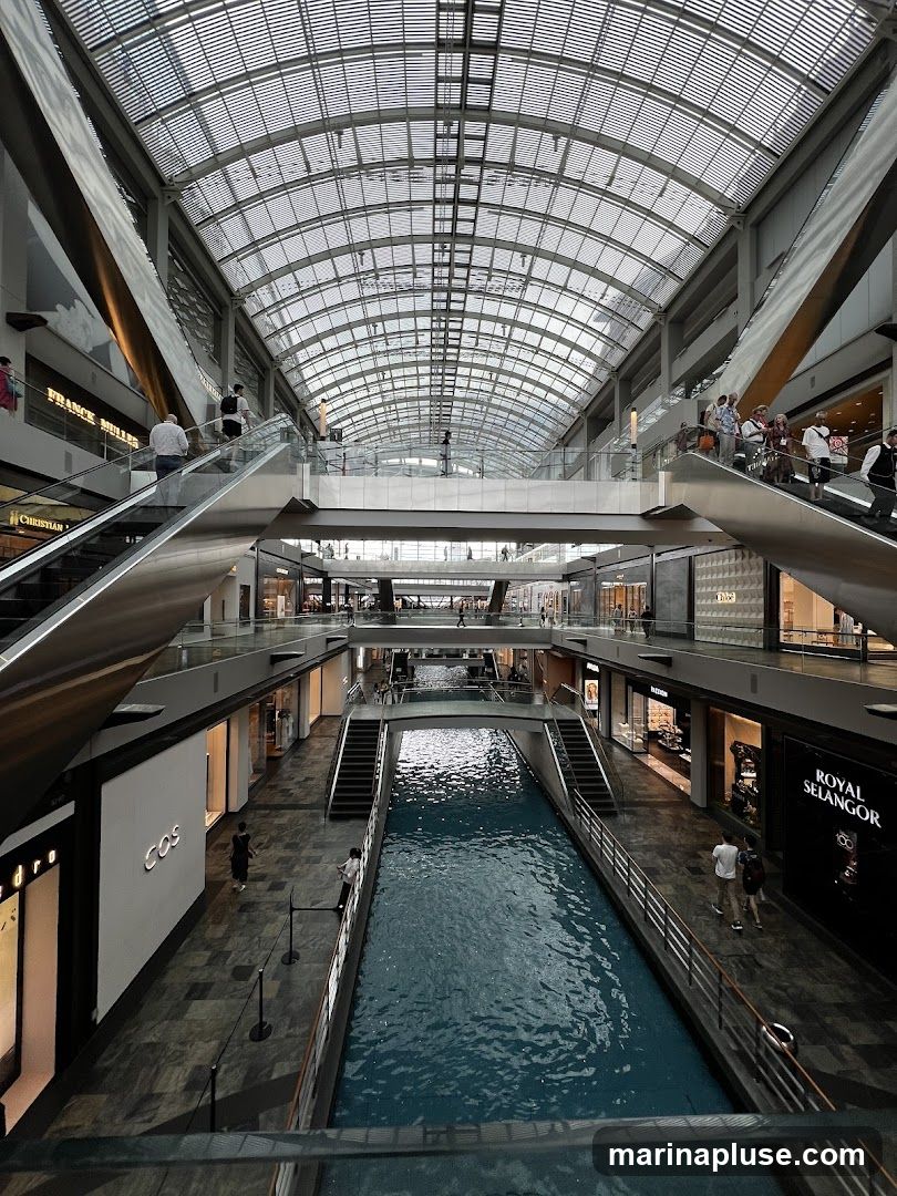 An interior view of a large, modern shopping mall featuring a canal running through its center. Multiple levels of shops and escalators are visible, with a glass ceiling allowing natural light to illuminate the space. People are seen walking, shopping, and using escalators. Storefronts for brands like COS and Royal Selangor are visible.