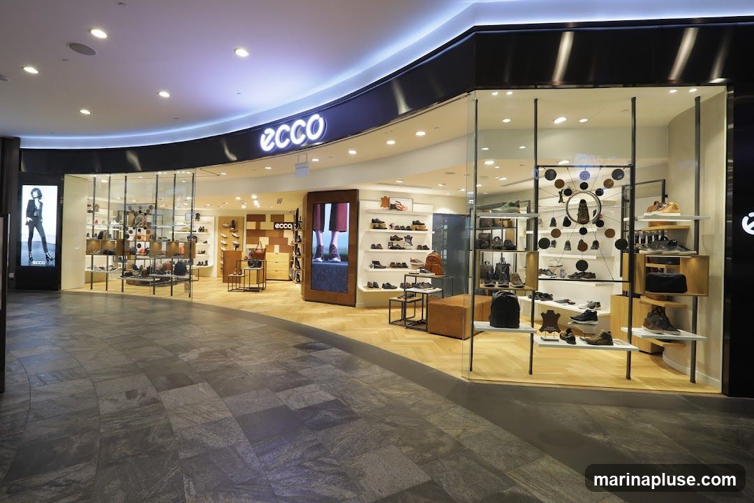 The exterior of an Ecco shoe store with a prominent "ecco" logo above the entrance. The store features large glass windows displaying various shoes and bags on shelves. Inside, the store is well-lit with modern shelving and wooden flooring. A digital display on the left shows a model wearing Ecco shoes.