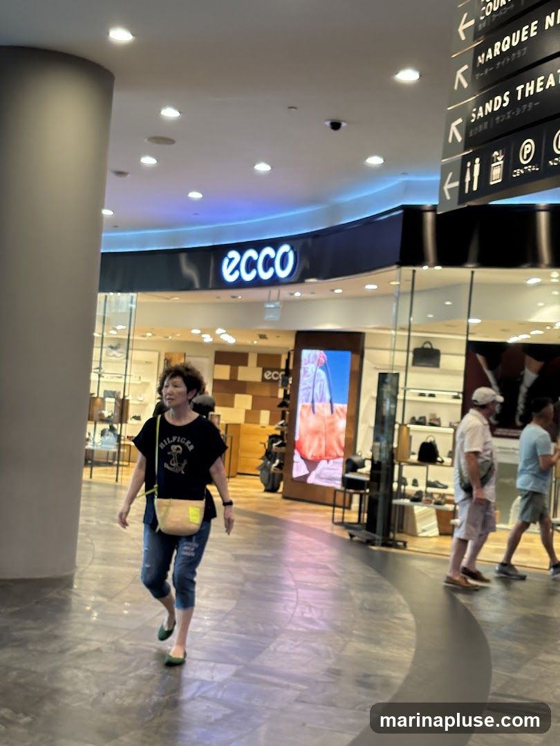 A woman walks past an Ecco store in a mall. The store has a large illuminated sign with the Ecco logo. Inside, shoes and bags are displayed on shelves. A digital screen shows an advertisement for clothing. Other shoppers are visible in the background.
