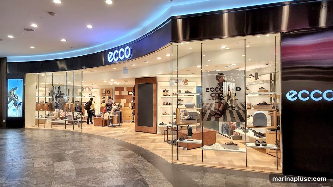 The exterior of an Ecco shoe store with large glass windows displaying shoes and accessories. The Ecco logo is visible on the facade and on a digital display. Several customers are browsing inside.