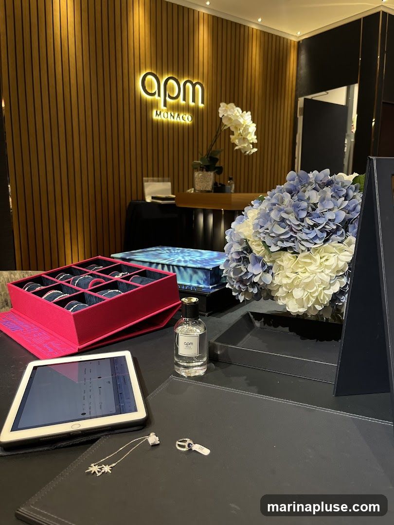A display table in a store features jewelry, a bottle of perfume, and a tablet. The background shows a wall with vertical wooden slats and a prominent illuminated "apm MONACO" logo. A bouquet of blue and white hydrangeas is placed on the right side of the table. In the foreground, a tablet displays what appears to be a website or catalog, alongside a necklace and a ring. A pink box filled with bracelets is also visible.