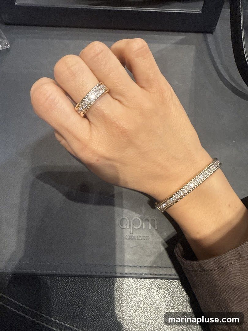 A hand wearing a diamond-encrusted ring and a matching bracelet. The jewelry is displayed on a dark surface with the APM Monaco logo visible.
