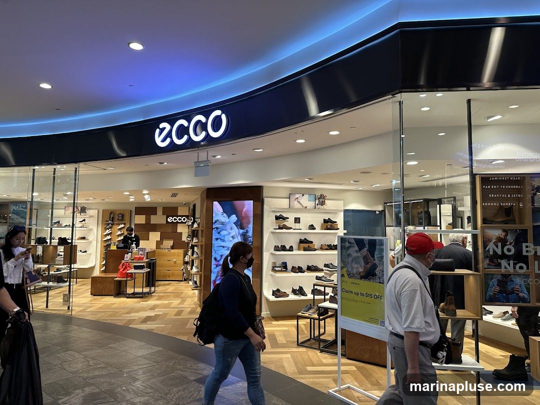 The exterior of an Ecco shoe store with the brand name prominently displayed above the entrance. The store features large glass windows showcasing various shoe displays and mannequins. Inside, shelves are stocked with shoes, and a cashier counter is visible. Several people are walking past the storefront.
