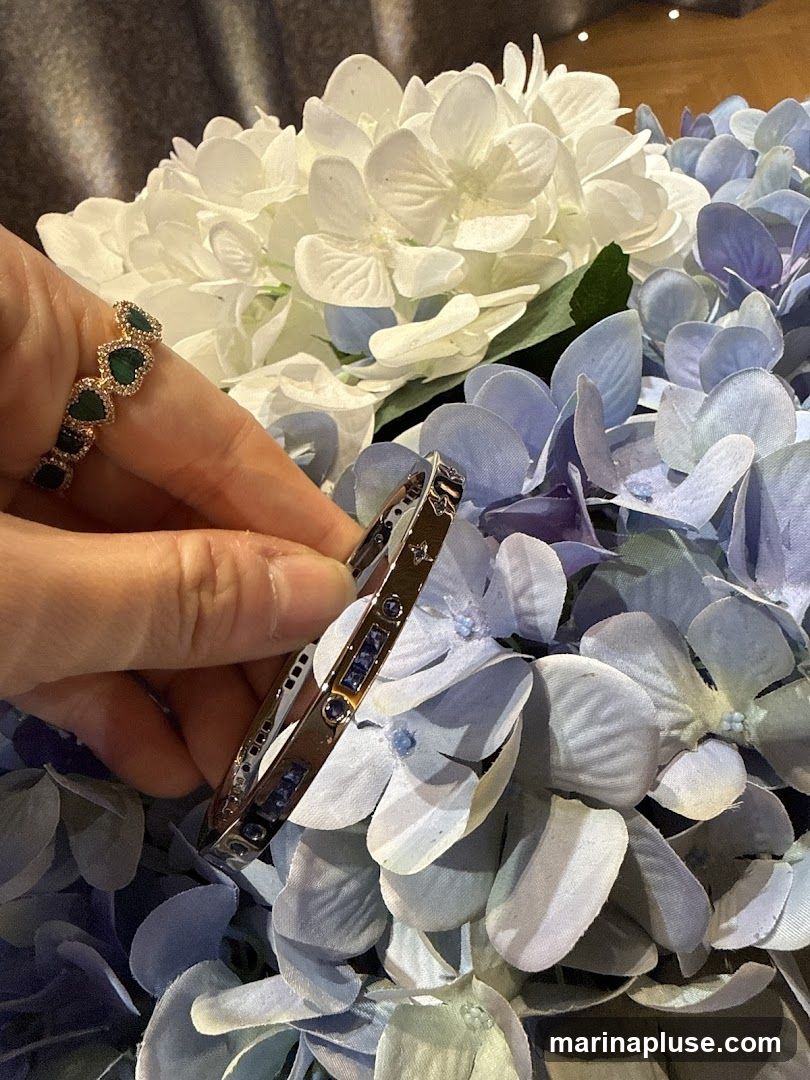 A hand holds a silver bangle bracelet with blue stones and a ring with green stones, set against a backdrop of white and blue hydrangeas.