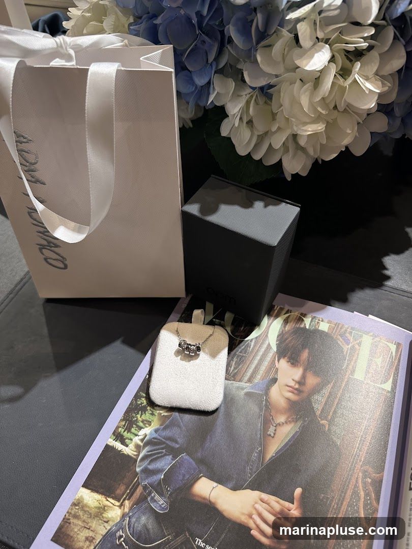A white shopping bag with ribbon handles and silver lettering, a dark grey jewelry box, a necklace displayed on a white stand, and a magazine featuring a man on the cover are arranged on a dark surface. A bouquet of blue and white hydrangeas is visible in the background.