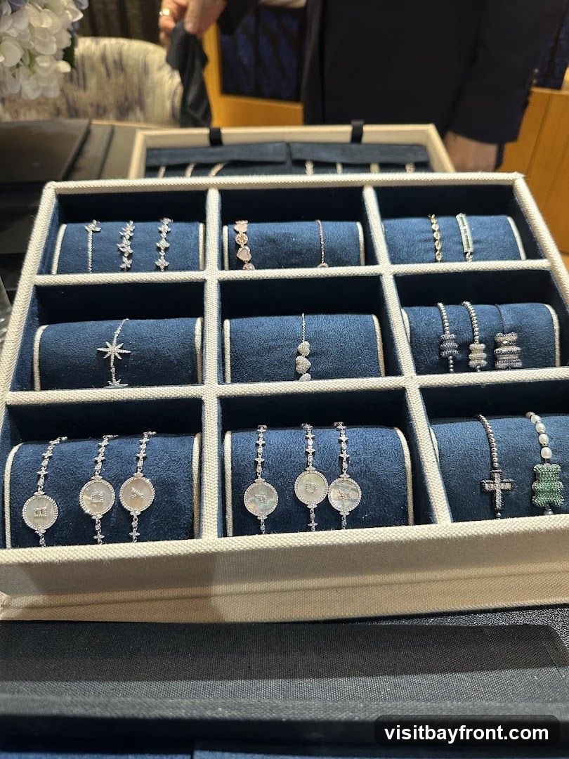 A close-up view of a jewelry display case filled with various bracelets. The bracelets are arranged in a grid of compartments, each resting on a dark blue velvet cushion. The jewelry includes delicate chains with small charms, some featuring celestial motifs like stars, others with circular pendants, and some with gemstone embellishments. One section shows bracelets with what appear to be zodiac symbols or monograms. Another section displays bracelets with crosses and a green bear charm. The overall presentation suggests a luxury jewelry store or a high-end boutique.