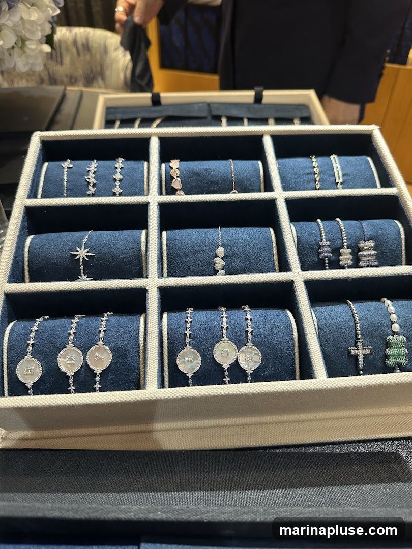 A close-up view of a jewelry display case filled with various bracelets. The bracelets are arranged in a grid of compartments, each resting on a dark blue velvet cushion. The jewelry includes delicate chains with small charms, some featuring celestial motifs like stars, others with circular pendants, and some with gemstone embellishments. One section shows bracelets with what appear to be zodiac symbols or monograms. Another section displays bracelets with crosses and a green bear charm. The overall presentation suggests a luxury jewelry store or a high-end boutique.