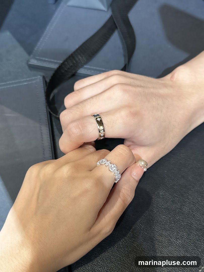 A close-up shot of two people's hands, adorned with rings, resting on a dark surface. The left hand, belonging to a woman, wears a delicate band encrusted with small, sparkling stones. The right hand, belonging to a man, displays a darker band with star-like motifs and small gems. In the background, a dark, textured bag or box is visible.