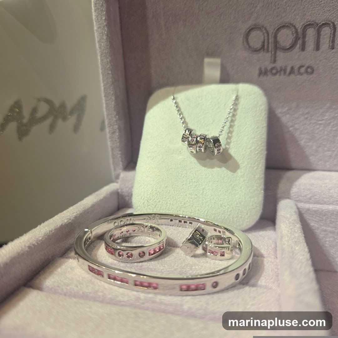 A silver necklace with a pendant of stacked rings, a silver bracelet with pink stones, and two silver rings with pink stones are displayed in a purple velvet jewelry box. The brand logo "apm MONACO" is visible on the inside lid of the box.