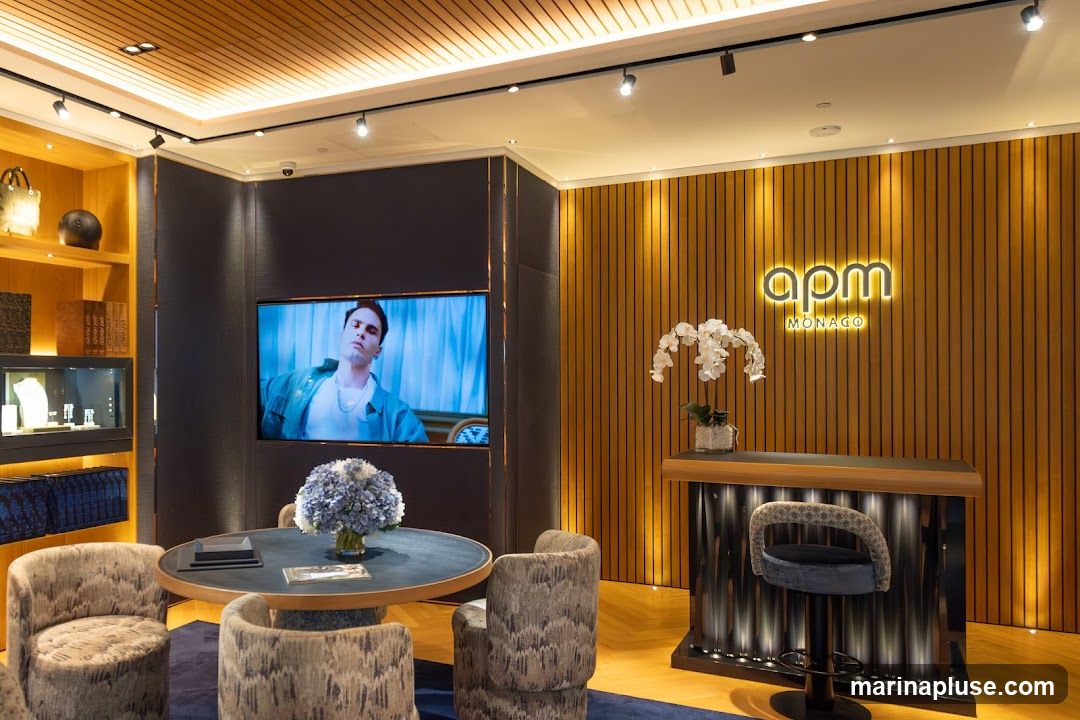 The interior of an APM Monaco store features a modern and luxurious design. A large screen displays a man's portrait. A round table with chairs is set up for seating. A counter with a stool is positioned against a wood-paneled wall, illuminated by a neon APM Monaco logo. Display shelves with jewelry and decorative items are visible on the left.