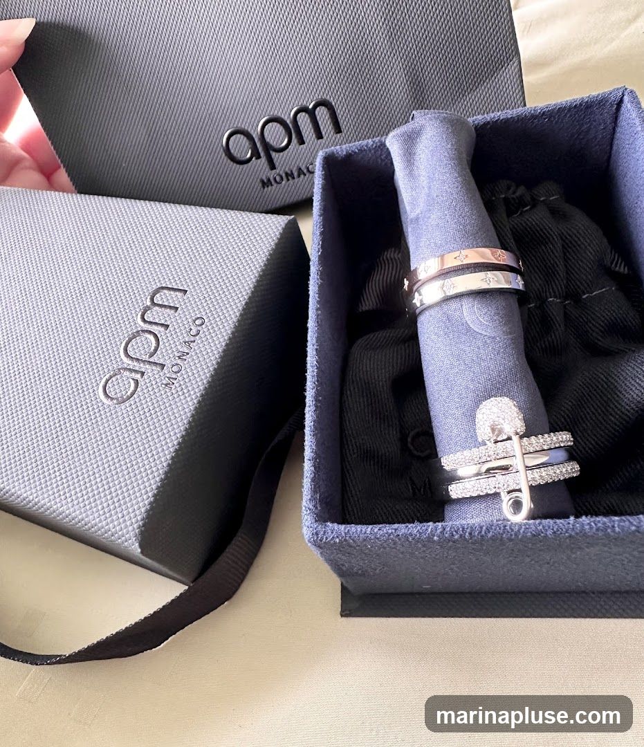 Two rings are displayed in a blue velvet box. One ring is rose gold with star patterns, and the other is silver with star patterns. A third ring, a silver band with a safety pin charm encrusted with small stones, is also in the box. Two branded boxes with the logo "apm MONACO" are visible in the background.