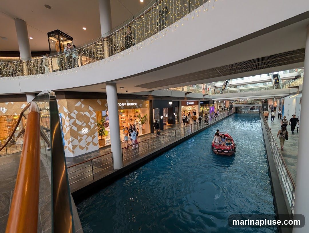A view of a shopping mall with a canal running through it. People are walking along the shops on the sides, and a small boat with passengers is on the water. The mall is decorated with string lights.