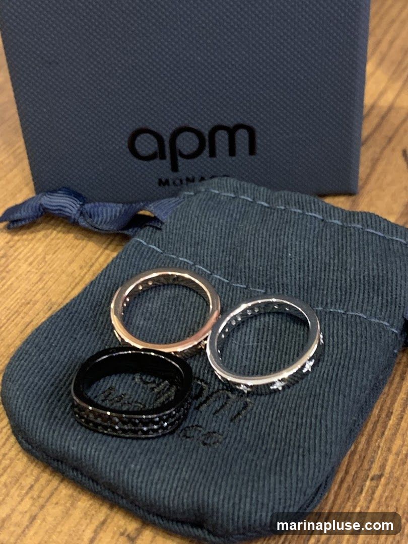 Three rings are displayed on a dark blue pouch. One ring is black and encrusted with small black stones. Another ring is rose gold and appears to be studded with small clear stones. The third ring is silver and has small star shapes around it. Behind the pouch is a dark blue box with the logo "apm MONACO" in black lettering. The items are placed on a wooden surface.