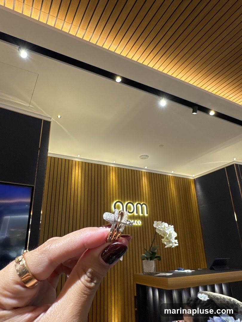 A hand holds up a diamond ring with a cross design in front of a store interior. The background features a wooden paneled wall with a glowing sign that reads "apm monaco". A white orchid sits on a counter to the right.