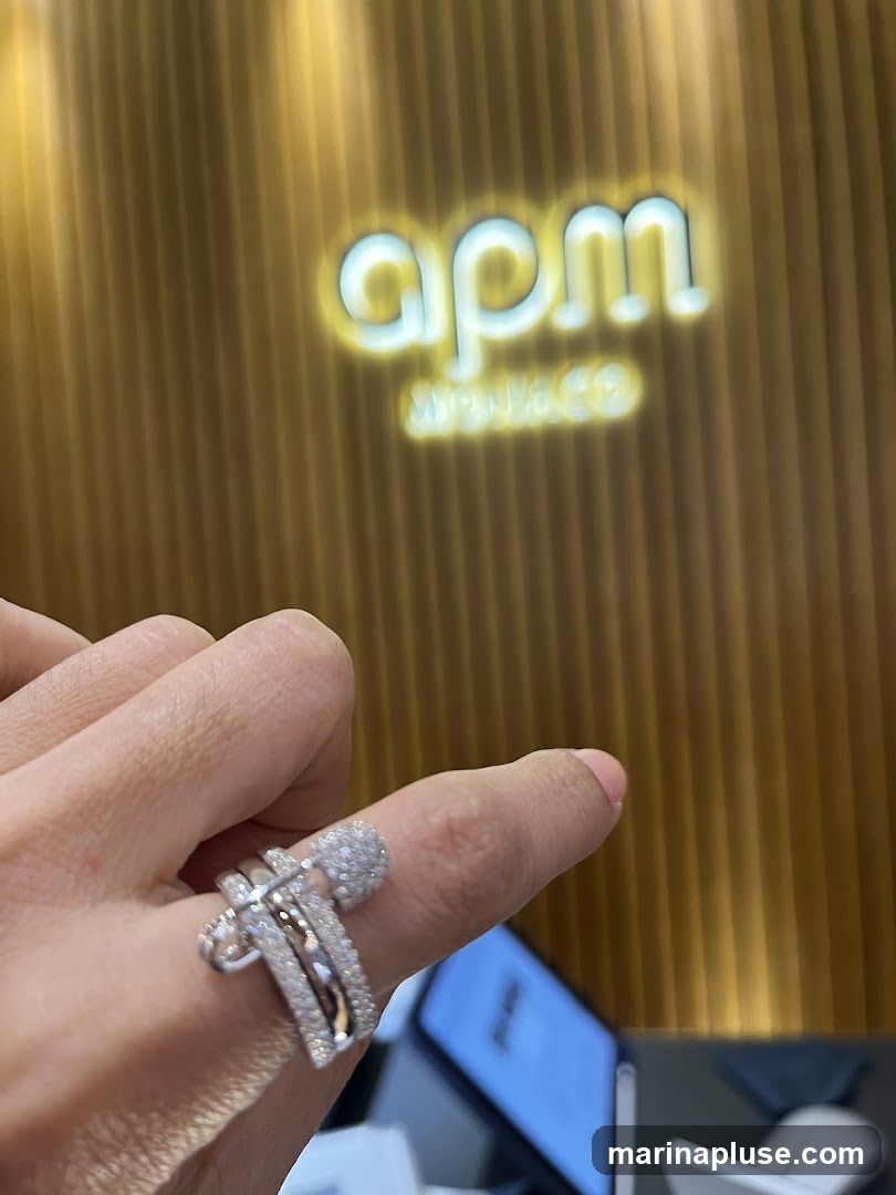 A close-up shot of a hand wearing a sparkling diamond ring. In the background, a blurred logo for 'apm monaco' is visible on a gold, ribbed wall.