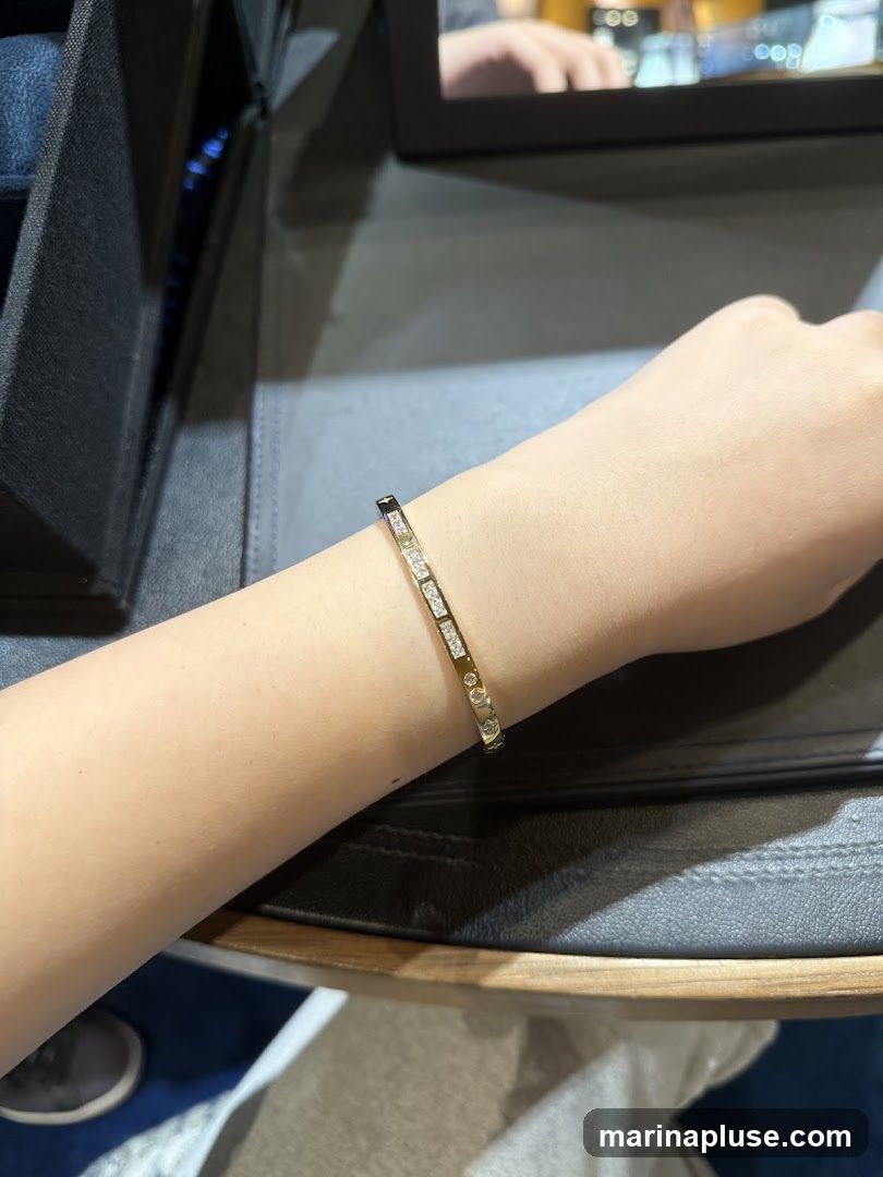 A close-up shot of a person's arm wearing a gold bangle bracelet adorned with diamonds. The bracelet is thin and features a repeating pattern of small square-set diamonds and round-cut diamonds. The background is blurred, showing a dark, textured surface, possibly a display case or table, and a reflection in a mirror or screen above.