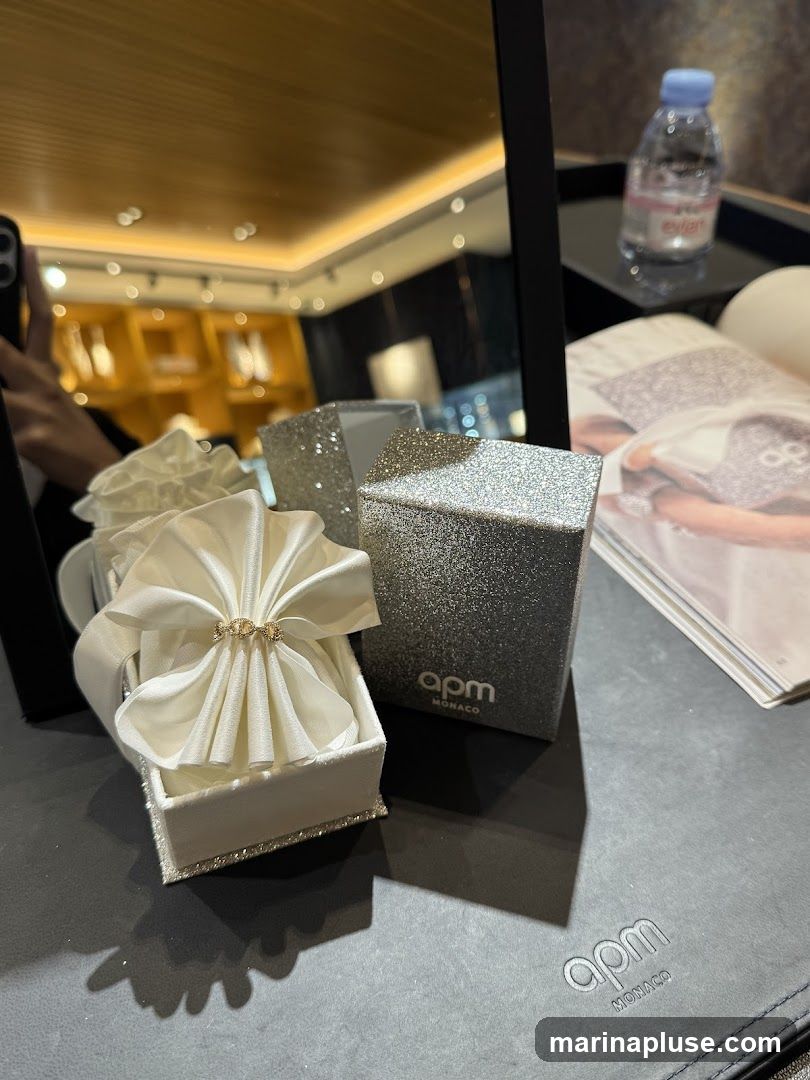 A white satin-lined jewelry box with a ring is displayed next to a glittery box with the APM Monaco logo. A magazine and a bottle of Evian water are also visible in the background.