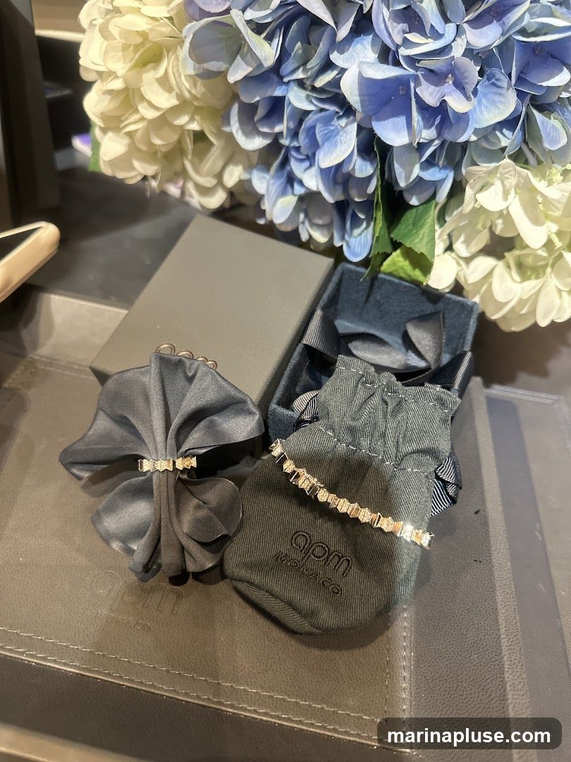 A close-up view of two pieces of jewelry, possibly bracelets, displayed on a dark textured surface. One piece is draped over a dark grey fabric, while the other is shown on a small drawstring pouch. Both pieces appear to be delicate and adorned with small, sparkling stones. The pouch has the logo "apm MONACO" printed on it. In the background, a bouquet of blue and white hydrangeas adds a touch of color and elegance to the scene. A grey gift box and a dark blue jewelry box are also visible.