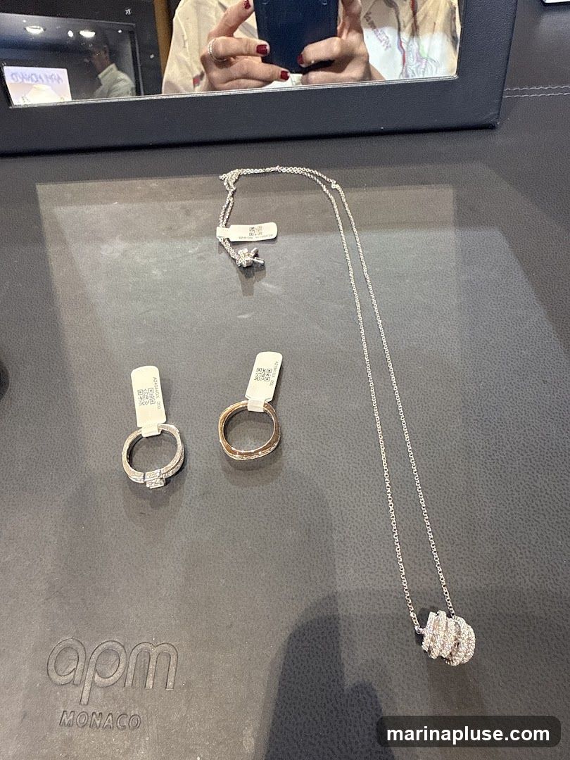 A display of jewelry on a dark surface, featuring two rings and a necklace. The rings appear to be a diamond engagement ring and a band. The necklace has a pendant with a coiled, diamond-encrusted design. Tags with QR codes are attached to the jewelry. The APM MONACO logo is visible on the surface. A reflection in a mirror above shows a person holding a phone and wearing jewelry.