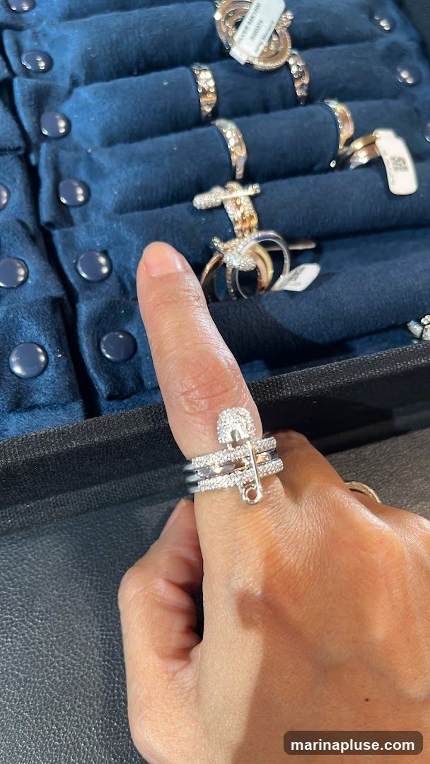 A hand wearing a diamond-encrusted safety pin ring. The ring is stacked with other diamond bands. In the background, a display case holds various rings on a dark blue velvet surface. Some rings have price tags attached.