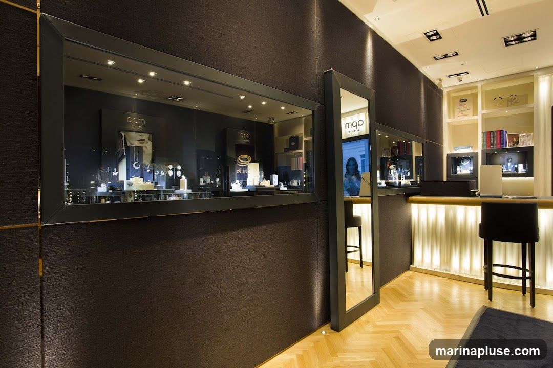The interior of a modern jewelry store with display cases showcasing necklaces, bracelets, and earrings. The walls are dark textured fabric, and the floor is polished wood. A counter with stools is visible, illuminated by soft lighting. A digital display shows a woman's face. Shelves behind the counter hold books and branded boxes.