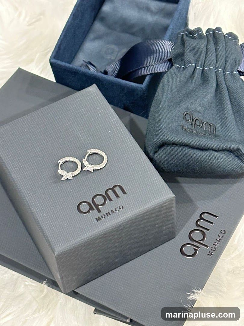 A pair of diamond hoop earrings rests on an open gray box with the APM Monaco logo. Beside it is a blue velvet jewelry box and a gray drawstring pouch, also branded with the APM Monaco logo.
