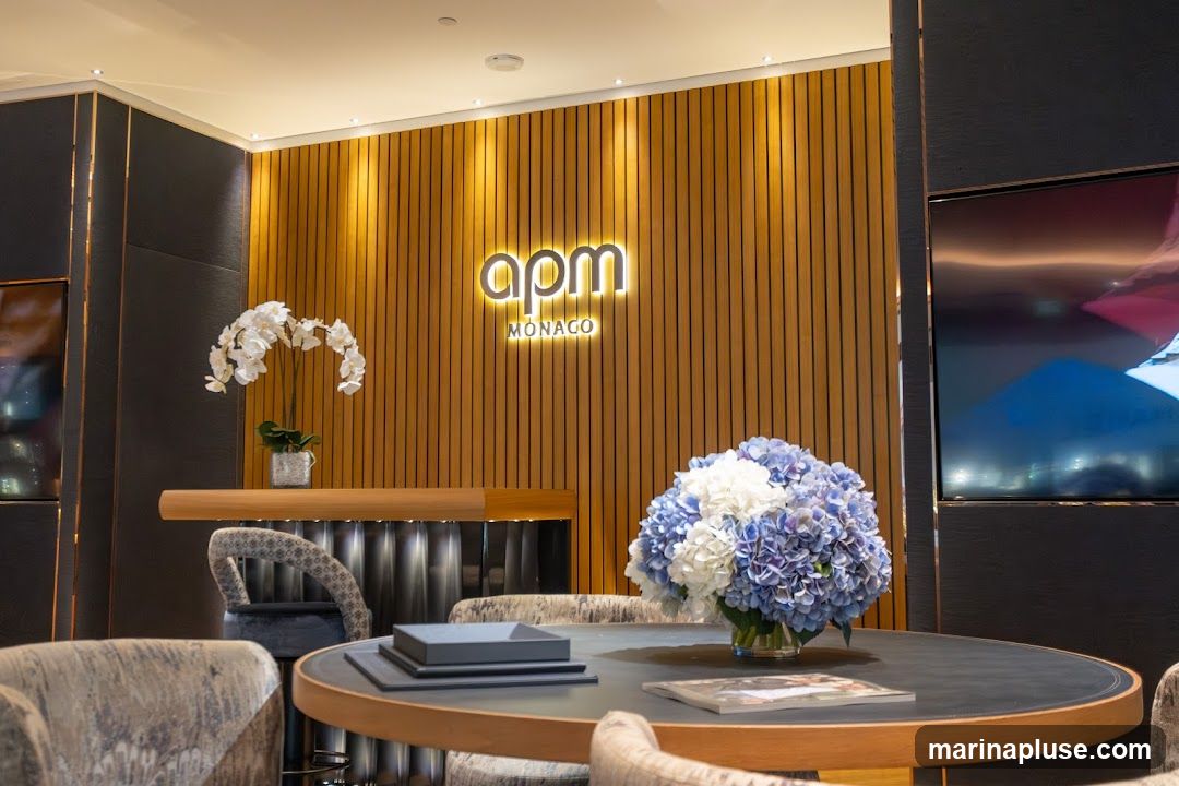 The interior of an APM Monaco store features a wooden slatted wall with the illuminated APM Monaco logo. A counter with chairs is visible, along with a round table adorned with a bouquet of blue and white hydrangeas and a magazine. Another bouquet of white orchids is placed on the counter. A large screen displays an abstract image.