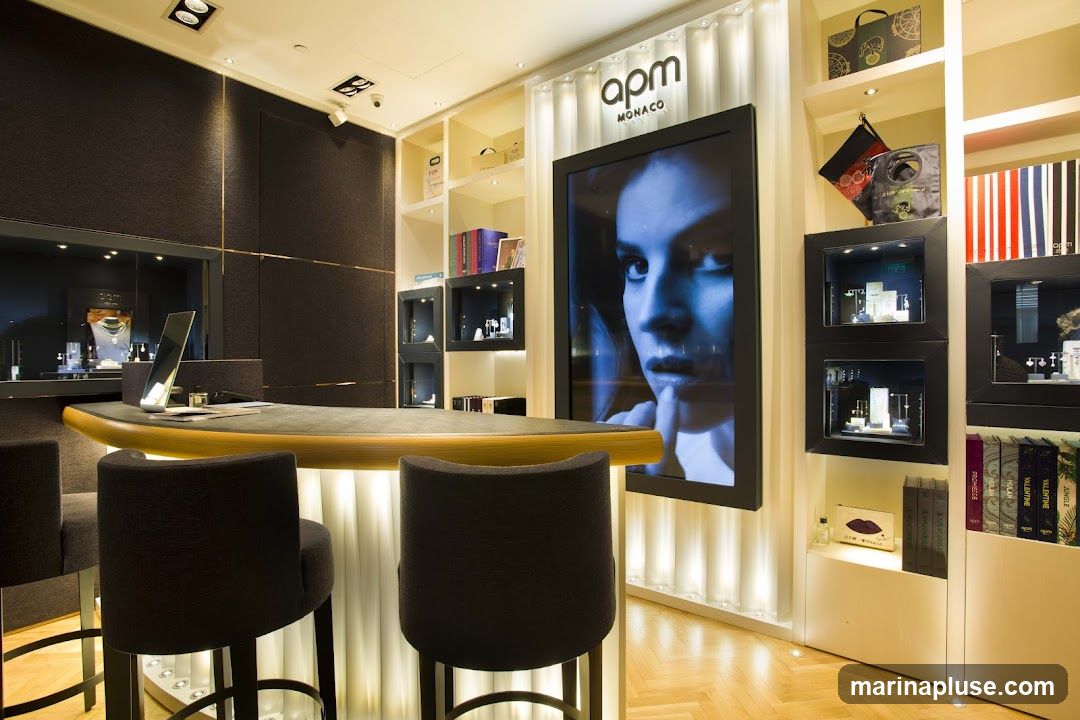The interior of a modern jewelry store with a curved counter, bar stools, and display cases. A large digital screen displays a woman's face. Shelves are stocked with branded bags and books. The "apm MONACO" logo is visible on the wall behind the screen.