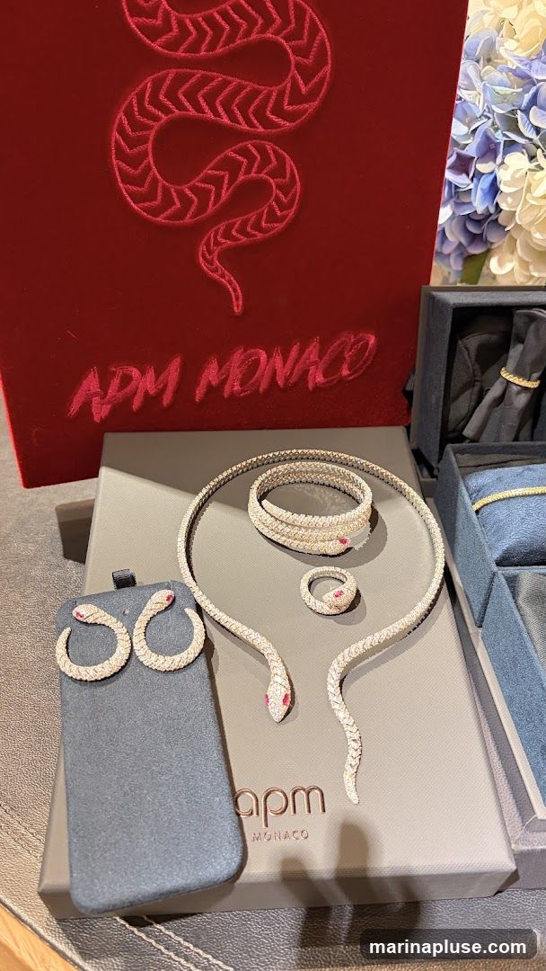 A display of snake-themed jewelry, including earrings, a necklace, a bracelet, and a ring, all encrusted with small stones. The jewelry is arranged on a gray surface with the brand name "apm MONACO" visible. Behind the jewelry is a red box with a red embroidered snake design and the text "APM MONACO" in cursive. To the right, a blue box contains a ring, and a bouquet of blue and white flowers is visible in the background.