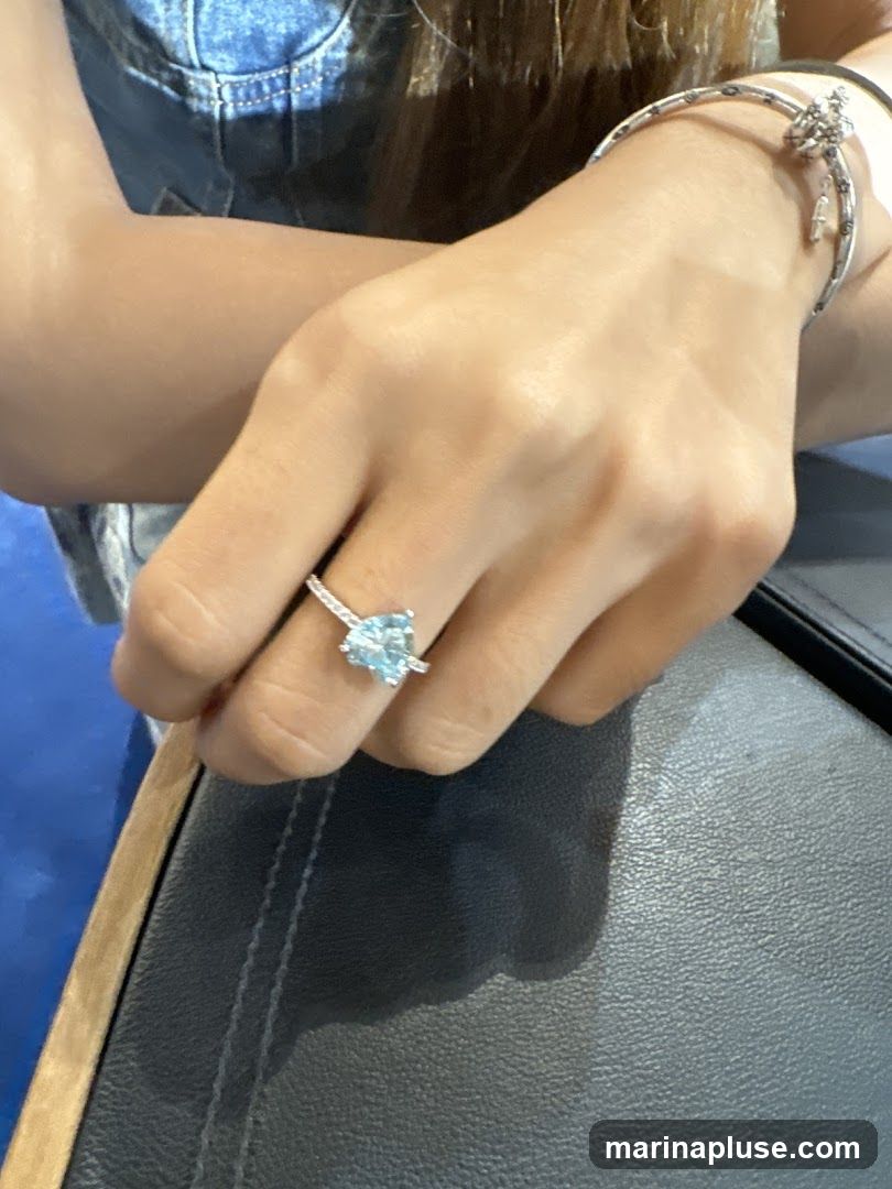 A close-up shot of a woman's hand wearing a heart-shaped aquamarine ring and a silver bracelet. She is wearing a denim top.