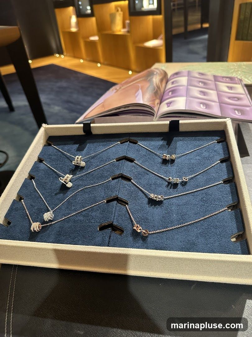 A display tray filled with delicate necklaces, each featuring a unique pendant. The necklaces are laid out on a dark blue velvet surface. In the background, an open magazine showcases images and text, and a well-lit display shelf with various items is visible.