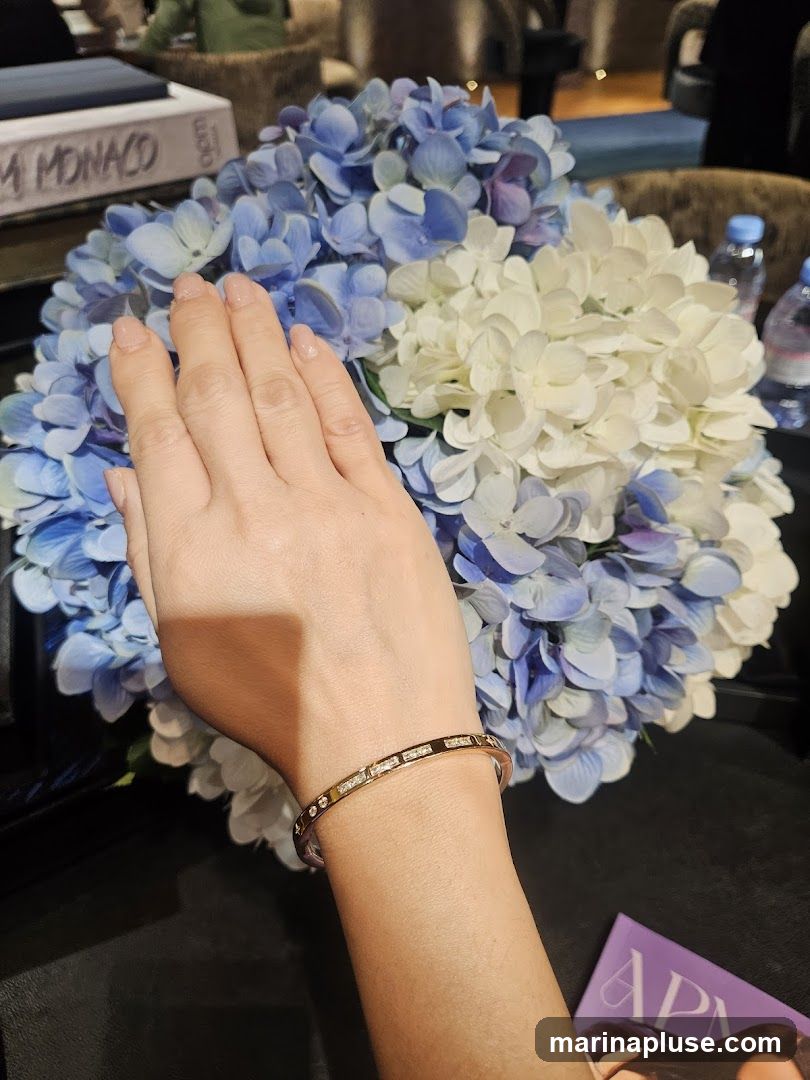 A close-up shot of a person's hand wearing a diamond-studded bracelet, positioned in front of a large bouquet of blue and white hydrangeas. In the background, a book with the text "MONACO" is visible, along with bottles of water and a magazine with the partial text "APM". The scene appears to be indoors, possibly in a lounge or hotel setting.