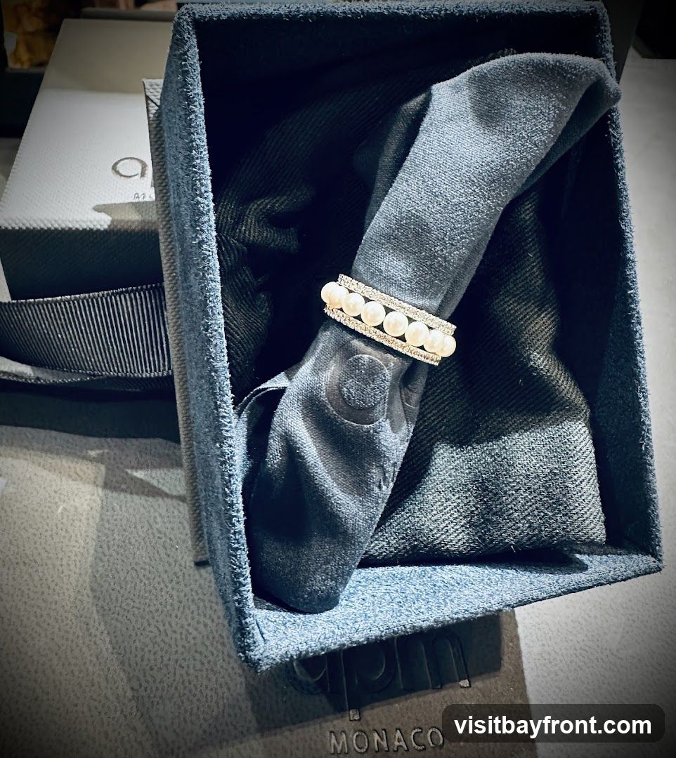 A close-up shot shows a ring with pearls and diamonds resting on a dark, folded cloth inside a blue velvet box. The box is open, revealing the ring and the cloth. A white box with a black ribbon and a logo is visible in the background. The word "MONACO" is visible on a surface below the box.