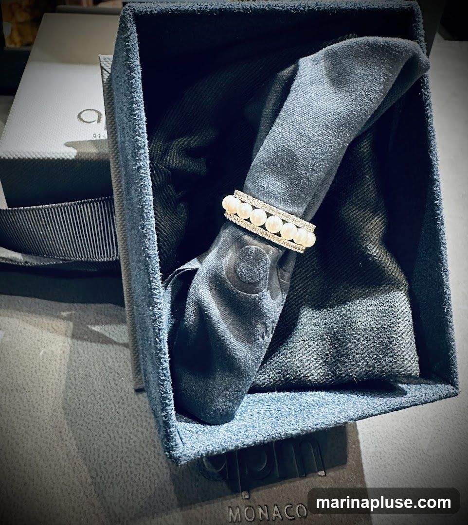 A close-up shot shows a ring with pearls and diamonds resting on a dark, folded cloth inside a blue velvet box. The box is open, revealing the ring and the cloth. A white box with a black ribbon and a logo is visible in the background. The word "MONACO" is visible on a surface below the box.
