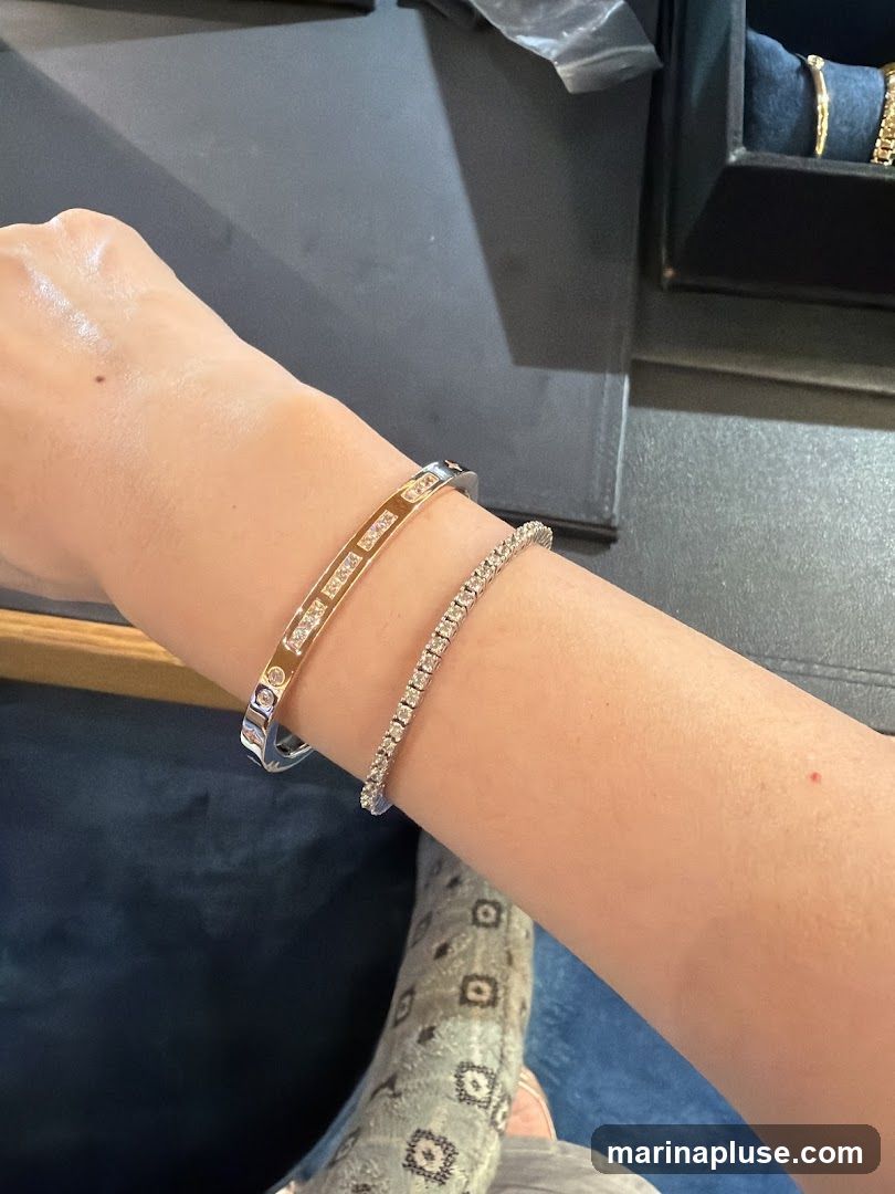 A person's arm is adorned with two bracelets. The upper bracelet is a silver-toned bangle with rectangular inlays, some of which appear to be diamonds. The lower bracelet is a delicate tennis bracelet, entirely composed of small, sparkling diamonds. In the background, a black jewelry box is visible, containing another gold bracelet.