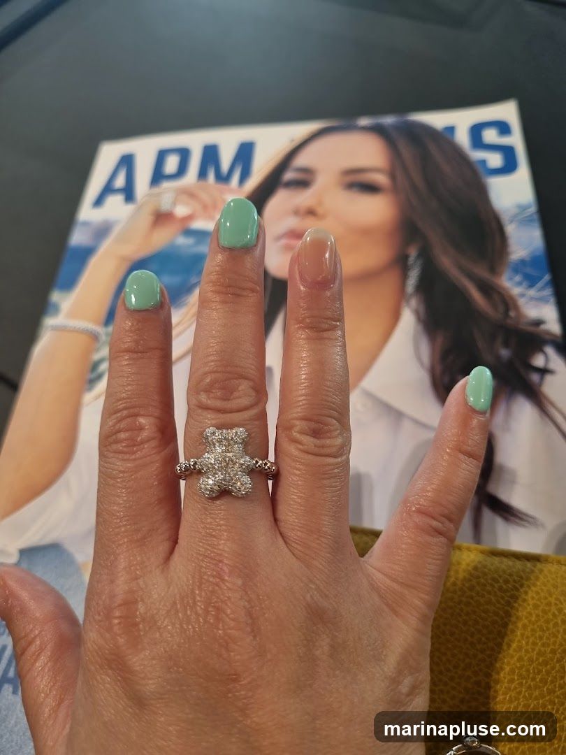 A woman's hand with a diamond-encrusted teddy bear ring and mint green nail polish is shown in the foreground. Behind the hand is a magazine cover featuring a woman with the text "APM" visible.
