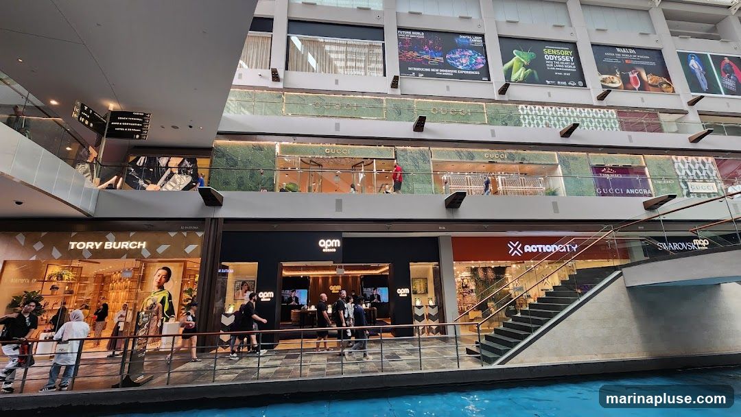 A wide shot of the interior of a multi-level shopping mall with various stores visible. On the lower level, the storefronts for Tory Burch, APM Monaco, ActionCity, and Swarovski are prominent. Above, the Gucci store is visible on the second level, with large advertisements on the upper levels for attractions like "Future World" and "Sensory Odyssey." A canal or water feature runs along the bottom of the image.