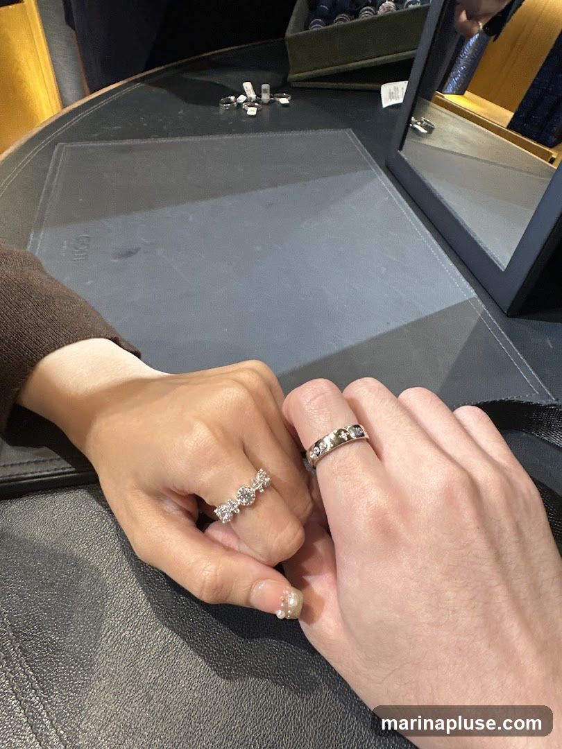 Two people holding hands, each wearing a ring. The person on the left is wearing a ring with multiple clear stones. The person on the right is wearing a band with small, dark stones. They are sitting at a dark, textured table, possibly in a jewelry store. A mirror is visible in the background, reflecting a display of jewelry.