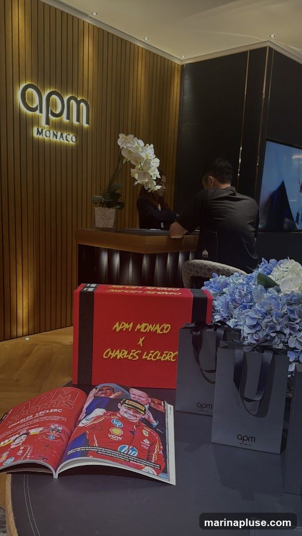 A display featuring APM Monaco branding, including a large red box with "APM MONACO X CHARLES LECLERC" text, an open magazine with images of Charles Leclerc, and several shopping bags with the APM Monaco logo. The background shows a reception desk with staff and a wall with the APM Monaco logo illuminated.