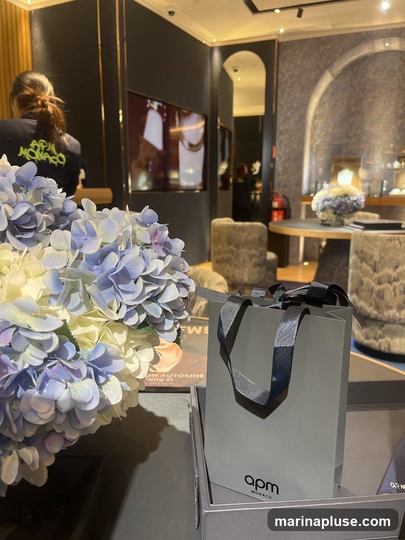A close-up view of a grey shopping bag with the APM Monaco logo, placed on a tray. In the foreground, a bouquet of blue and white hydrangeas adds a touch of color. The background reveals the interior of a jewelry store, with display cases, seating, and a television screen showing jewelry. A person wearing a shirt with the APM Monaco logo is visible in the left background.