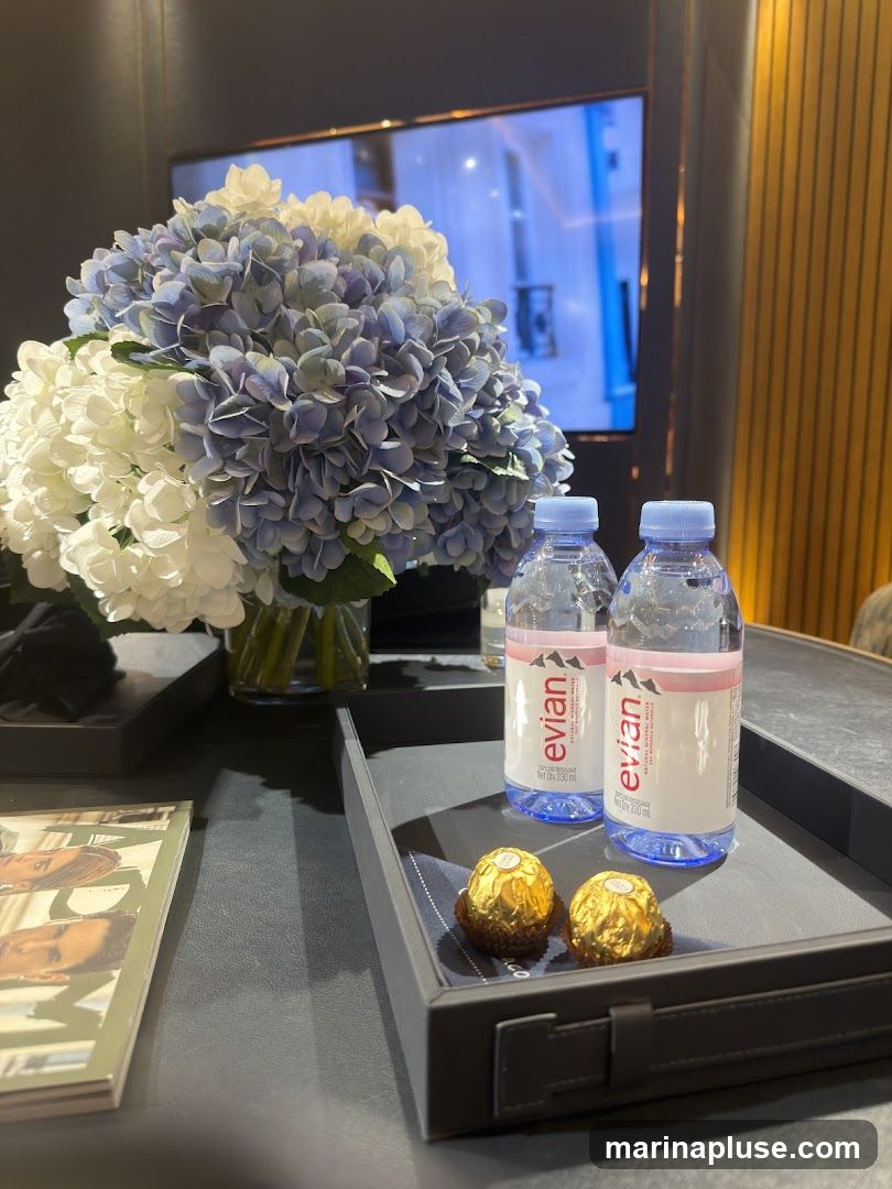 A dark tray holds two bottles of Evian water and two Ferrero Rocher chocolates. A vase of blue and white hydrangeas sits to the left, and a magazine is partially visible in the foreground. A television screen is in the background.