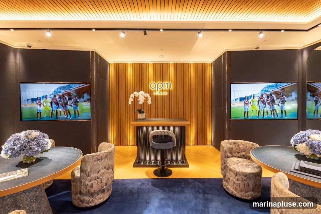 The image shows a well-lit, modern retail interior. In the center, a reception desk with the logo "apm MONACO" is visible, adorned with a white orchid. Two large screens on either side display images of a sports team. In the foreground, there are two round tables with floral arrangements and comfortable seating areas. The flooring is a light-colored wood herringbone pattern, and a dark blue carpet covers the central area.