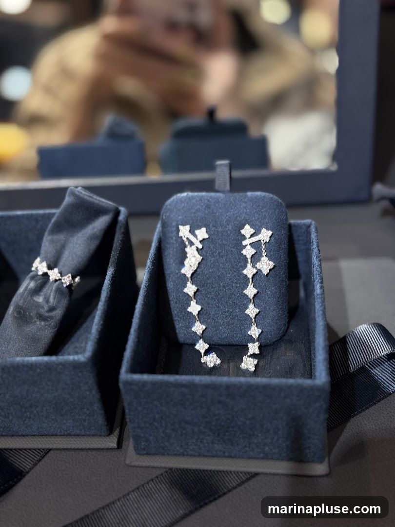 A close-up shot of diamond earrings and a ring displayed in dark blue velvet jewelry boxes. The earrings are long and dangle, featuring a pattern of small diamond-encrusted shapes. The ring, partially visible in its box, also appears to be set with diamonds. In the blurred background, a reflection shows a person holding a phone, possibly taking the photo, and display shelves.